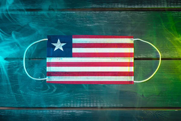 Medical mask for protection against airborne diseases, painted in the national flag of Liberia on a black background in blue-green smoke. Medical protection against airborne diseases, coronavirus