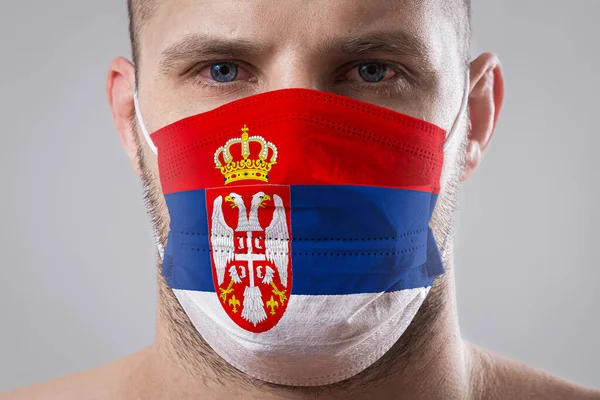 Young man with sore eyes in a medical mask painted in the colors of the national flag of Serbia. Medical protection against airborne diseases, coronavirus. Man is afraid of getting the flu