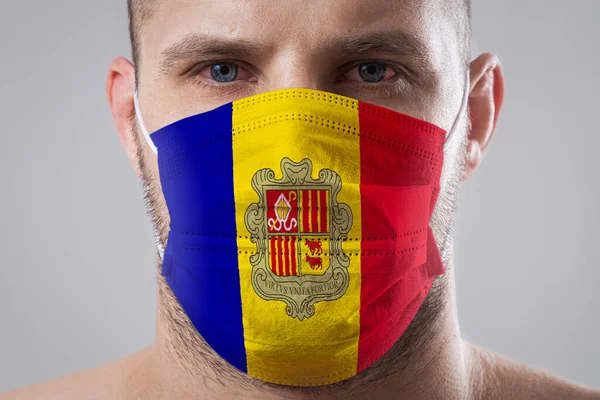 Young man with sore eyes in a medical mask painted in the colors of the national flag of Andorra. Medical protection against airborne diseases, coronavirus. Man is afraid of getting the flu