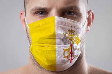 Vatikan bayrağının renklerine boyanmış tıbbi maskeli, yaralı gözlü genç bir adam. Hava hastalıklarına karşı tıbbi koruma, koronavirüs. Adam grip olmaktan korkuyor.