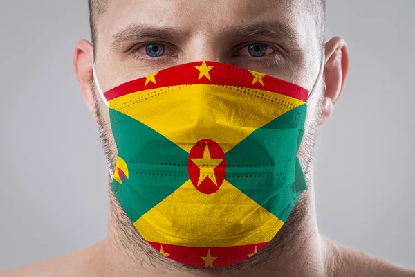 Young man with sore eyes in a medical mask painted in the colors of the national flag of Grenada. Medical protection against airborne diseases, coronavirus. Man is afraid of getting the flu