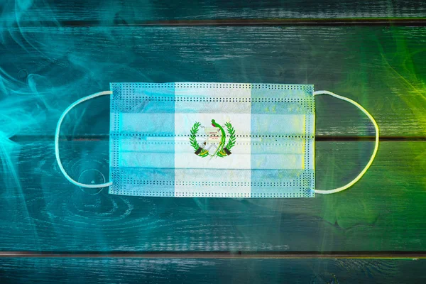 Medical mask for protection against airborne diseases, painted in the national flag of Guatemala on a black background in blue-green smoke. Medical protection against airborne diseases, coronavirus