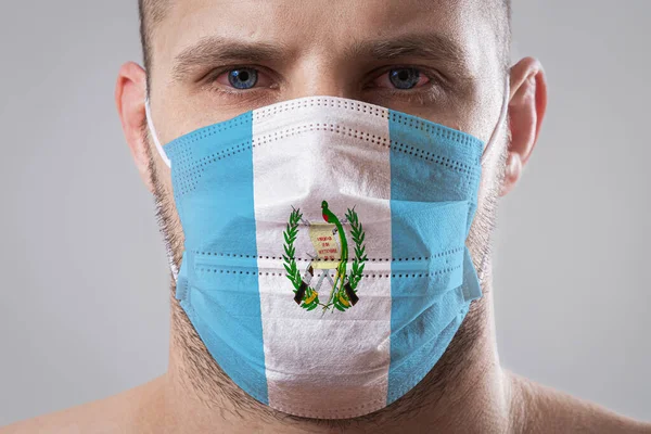 Young man with sore eyes in a medical mask painted in the colors of the national flag of Guatemala. Medical protection against airborne diseases, coronavirus. Man is afraid of getting the flu