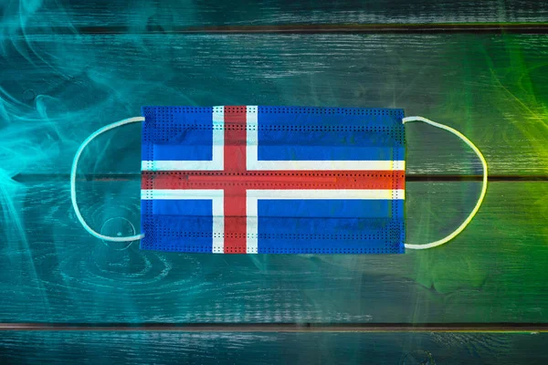 Medical mask for protection against airborne diseases, painted in the national flag of Iceland on a black background in blue-green smoke. Medical protection against airborne diseases, coronavirus