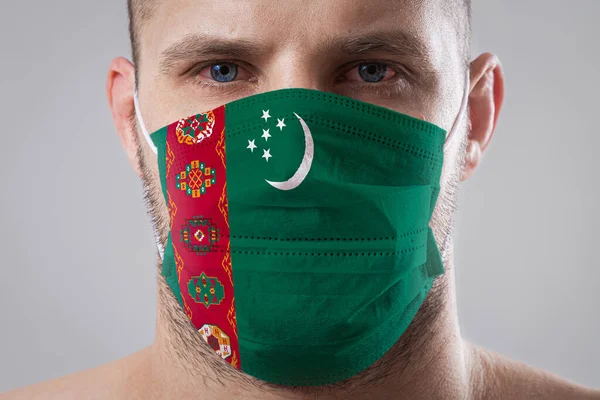 Young man with sore eyes in a medical mask painted in the colors of the national flag of Turkmenistan. Medical protection against airborne diseases, coronavirus. Man is afraid of getting the flu