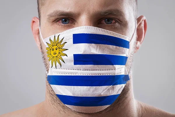 Young man with sore eyes in a medical mask painted in the colors of the national flag of Urugua
