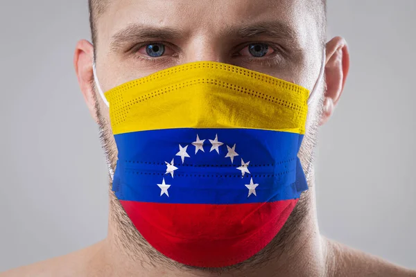 Young man with sore eyes in a medical mask painted in the colors of the national flag of Venezuela. Medical protection against airborne diseases, coronavirus. Man is afraid of getting the flu