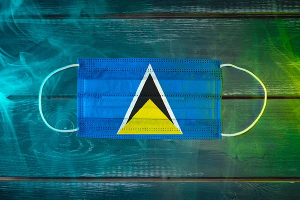 Medical mask for protection against airborne diseases, painted in the national flag of Saint Lucia on a black background in blue-green smoke. Medical protection against airborne diseases, coronavirus