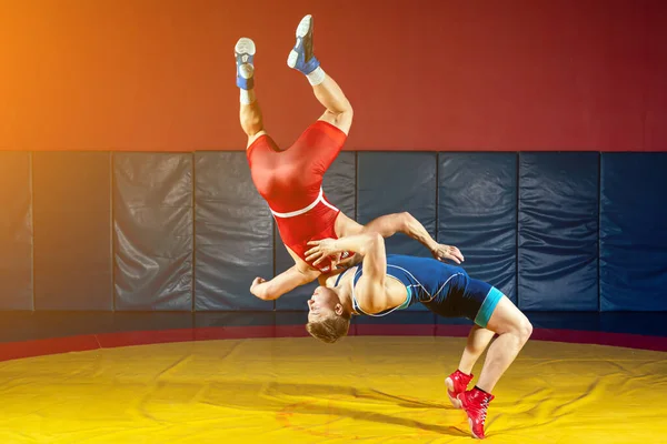 两名身穿蓝色和红色摔跤紧身衣的强壮男子正在体育馆的一条黄色摔跤地毯上摔跤和进行杂技摔跤 摔角手手忙脚乱 — 图库照片