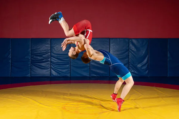 青と赤のレスリングのタイツを着た2人の強い男が格闘し ジムで黄色のレスリングカーペットの上で絶妙なレスリングをしています グラップルをやっている力士 — ストック写真