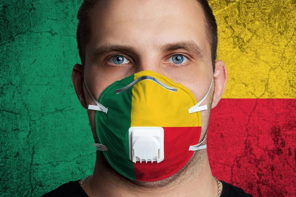 Young man with sore eyes in a medical mask painted in the colors of the national flag of Benin. Coronovirus disease  COVID-19 concept.  Man is afraid of getting the flu
