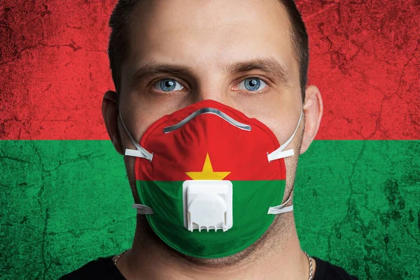 Young man with sore eyes in a medical mask painted in the colors of the national flag of Burkino Faso. Coronovirus disease  COVID-19 concept.  Man is afraid of getting the flu