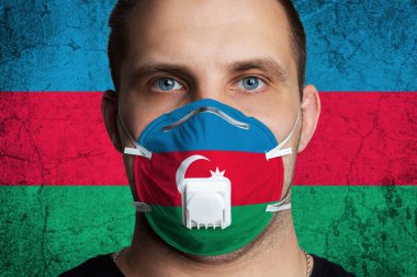 Azerbaycan bayrağının renklerine boyanmış tıbbi maskeli yaralı gözlü genç bir adam. Coronovirus hastalığı COVID-19 konsepti. Adam grip olmaktan korkuyor.