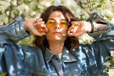 Çiçekli yaratıcı Portre Kadını. Tasarım sanat konsepti. Kıvırcık saçlı, kot ceketli, şık sarı gözlüklü, çiçek açan elma ağacı deresinin arka planında genç bir kadının romantik portresi. 90 biçimli portre 