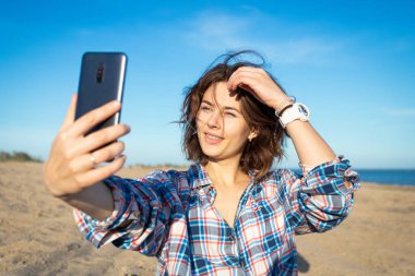 Koyu renk saçlı neşeli bir kadın gülümser, telefonda selfie çeker, plaj boyunca yürür, yaz günlerinde parlak güneşin tadını çıkarır. Denizde yaz tatili ve yaşam tarzı kavramı