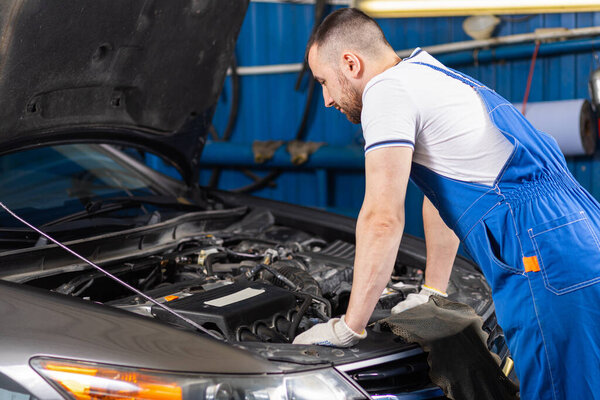 Handsome young male auto mechanic in special uniform clothes  looking for breakdown and repairing under the hood in the car engine in a car workshop