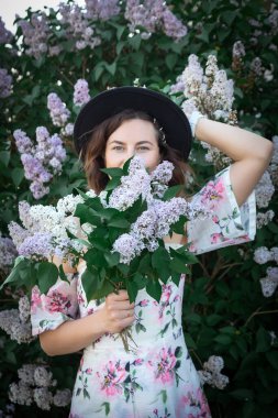 Yazlık çiçekli elbise giyen oldukça taze koyu renk saçlı genç bir kadın çiçek açan leylak ağacının arka planına poz veriyor. Köyde yaz tatili ve yaşam tarzı kavramı. Çiçekli genç bir kadın portresi