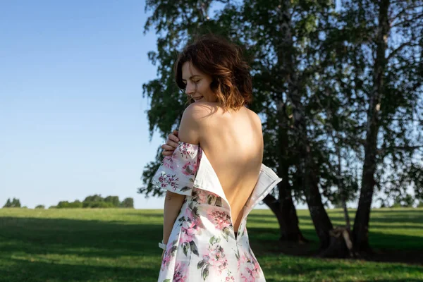 A beauty attractive woman in a light dress with bare shoulders and a floral print stands with her back to the camera with her dress zipped open. The concept of loneliness, thoughtfulness and longing for a woman. Close-up of a female bare back