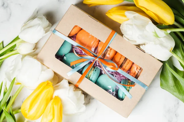 Box with orange and turquoise macaroons sweeties on marble table with bouquet of white and yellow tulips. Holiday concept