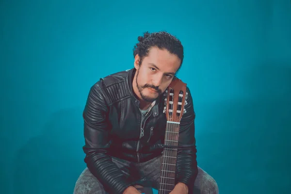 Porträt eines attraktiven Lateinamerikaners in Rockerjacke, auf blauem Hintergrund, mit Akustikgitarre — Stockfoto