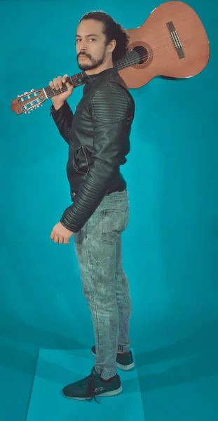 Привлекательный латиноамериканец в куртке рок-звезды, на синем фоне, показывающий акустическую гитару на камеру — стоковое фото