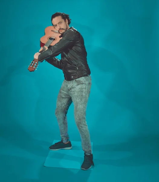 Attraente uomo latino in giacca rock star, su sfondo blu, prendendo la chitarra acustica come una mazza da baseball — Foto Stock
