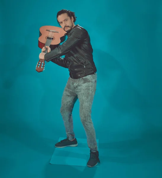 Attraktiver lateinischer Mann in Rockstar-Jacke, auf blauem Hintergrund, der die Akustikgitarre wie einen Baseballschläger nimmt — Stockfoto