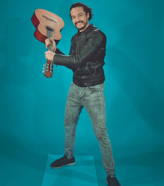 Atractivo hombre latino en chaqueta estrella de rock, sobre fondo azul, tomando la guitarra acústica como un bate de béisbol — Foto de Stock