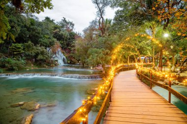 Huasteca Potosina 'daki Tamasopo Spa' nın panoramik manzarası, Şeffaf sular, bereketli bitki örtüsü, sarı ışıklarla aydınlatılmış eski ahşap köprü.