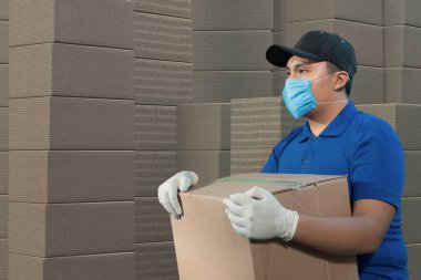 Ev teslimatı, online sipariş. Üniformalı bir adam, tıbbi bir maske ve kutulu lastik eldivenler elinde bir paket, kutularla dolu bir depoda.
