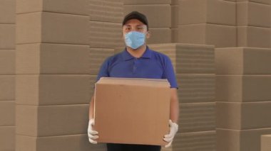 Ev teslimatı, online sipariş. Üniformalı bir adam, tıbbi bir maske ve kutulu lastik eldivenler elinde bir paket, kutularla dolu bir depoda.