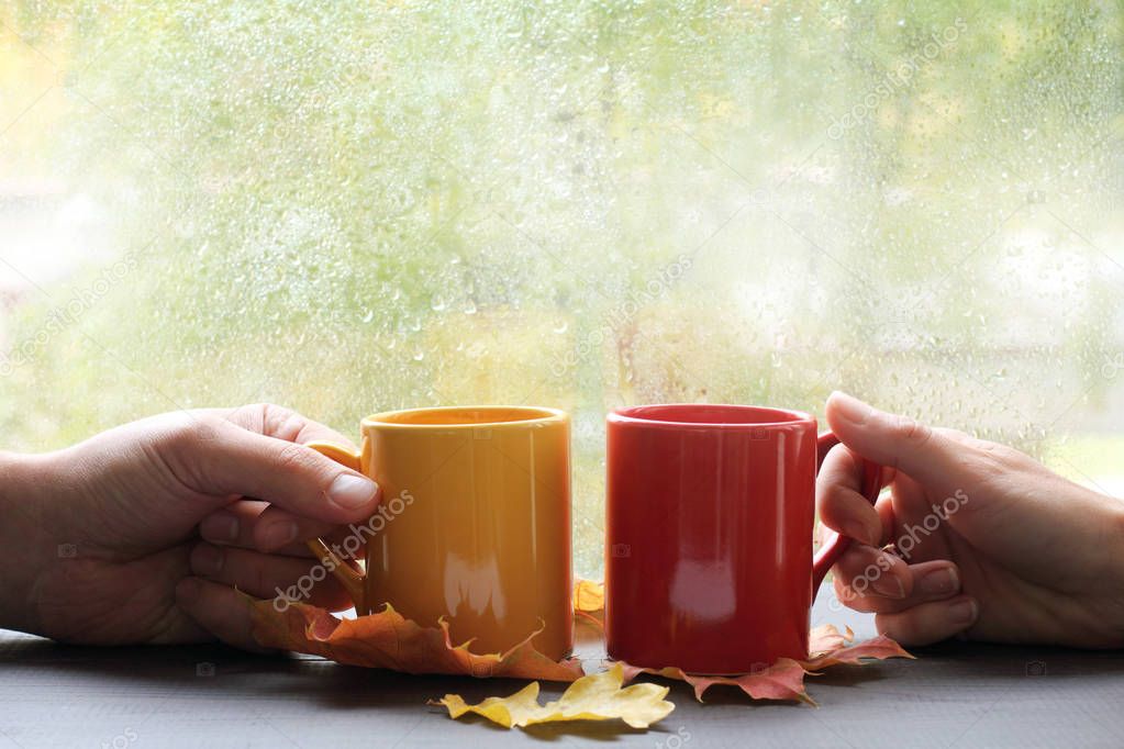 two hands holding cups on the background of wet window with raindrops / together tastier and warmer