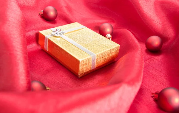 Золотая подарочная коробка с луком, на красной волнистой атласной ткани с рождественскими шарами . — стоковое фото