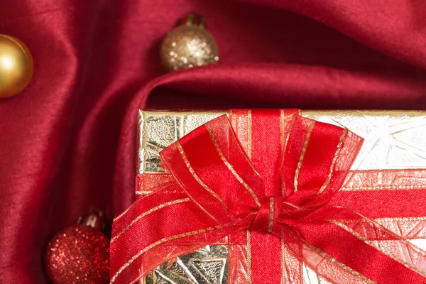 Confezione regalo con fiocco, su tessuto di raso ondulato rosso con palline di Natale, primo piano . — Foto Stock