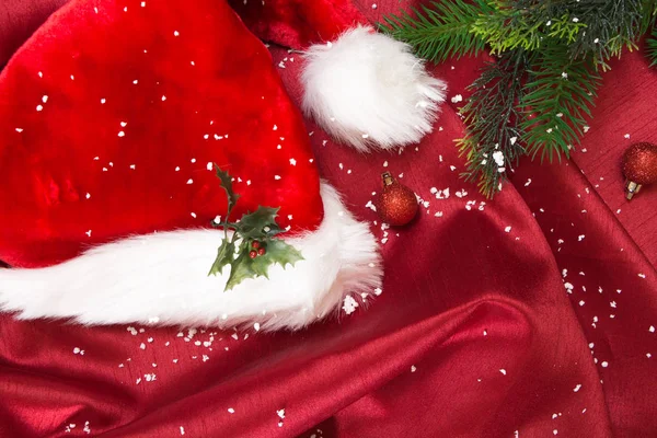 Cappello di Babbo Natale con ornamenti natalizi e neve, su tessuto ondulato rosso . — Foto Stock