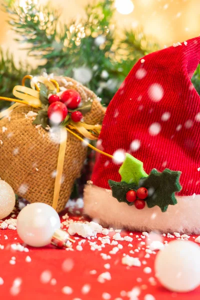 Cappello di Babbo Natale con ornamenti natalizi e neve che cade, su tessuto rosso . — Foto Stock