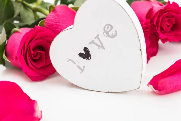 Bílé dřevěné srdce s slovem láska na ní a kyticí rudých růží a okvětních lístků — Stock fotografie