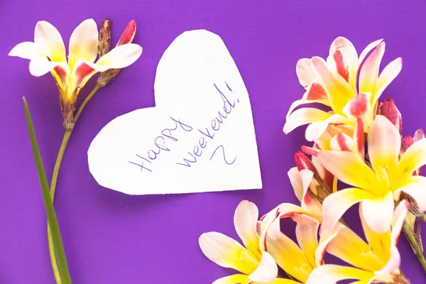 Nota en forma de corazón con palabras "¡Feliz fin de semana!" con flores en la superficie púrpura . —  Fotos de Stock