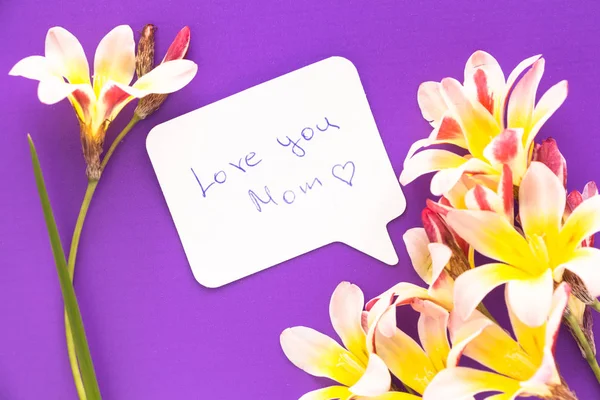 Note in shape of heart with words "Love you Mom!" with flowers on purple surface. — Stock Photo, Image