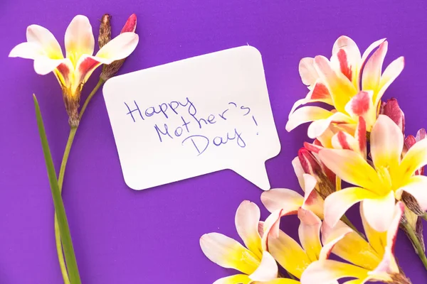 Nota en forma de corazón con palabras "¡Feliz Día de la Madre!" con flores en la superficie púrpura . —  Fotos de Stock