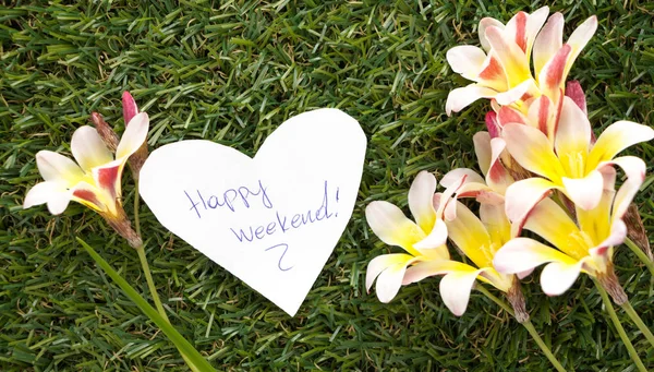 Note in shape of heart with words Happy Weekend!, with flowers on green grass. — Stock Photo, Image