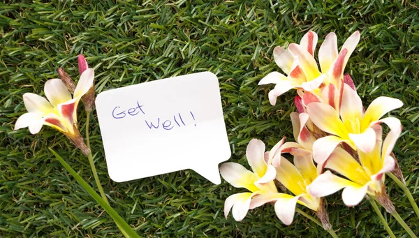 Note in shape of a chat bubble, with words Get Well! and flowers on green grass. — Stock Photo, Image