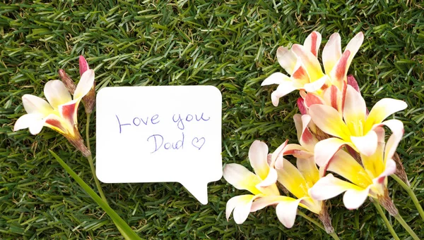 Note in shape of a chat bubble, with words Love you Dad! and flowers on green grass. — Stock Photo, Image