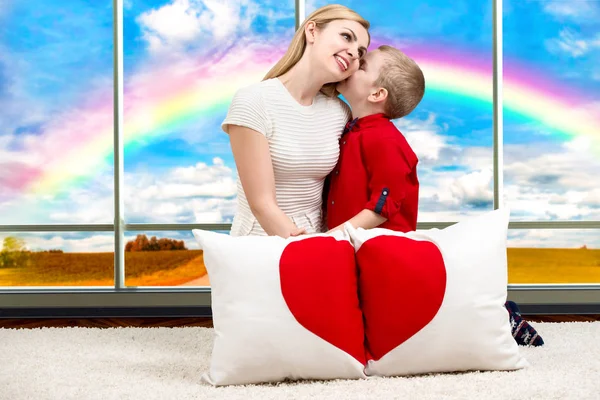 年轻的母亲带着美丽的宝贝儿子抱着枕头在心了彩虹般的全景窗口背景上的形状. — 图库照片