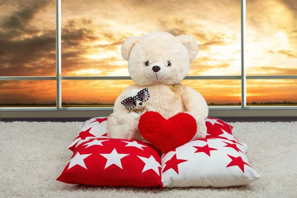 软软的艾德熊，戴着眼镜从太阳给了心，坐在枕头上的全景窗口背景. — 图库照片