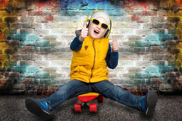 Niño sentado en el monopatín contra una pared de ladrillo.Moda infantil . — Foto de Stock