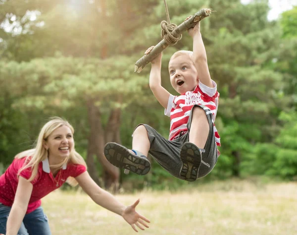 Kisfiú, lovaglás a rögtönzött swing az erdőben. Anyuka nézi a repülés, egy kis fia. — Stock Fotó
