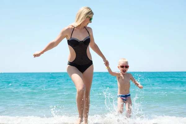 Mooie jonge moeder en kleine zoon hand in hand op de golven op het strand. Leuke, familie, vriendelijke zomervakantie. — Stockfoto
