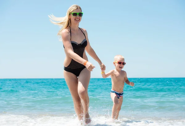 Mooie jonge moeder en kleine zoon hand in hand op de golven op het strand. Leuke, familie, vriendelijke zomervakantie. — Stockfoto