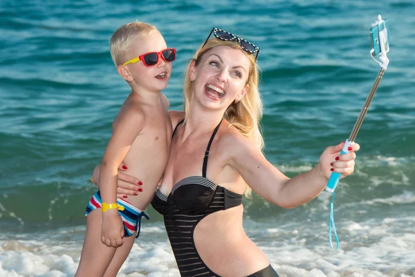 Madre e hijo en trajes de baño y gafas de sol tomando una selfie en un teléfono móvil en la costa del mar . —  Fotos de Stock